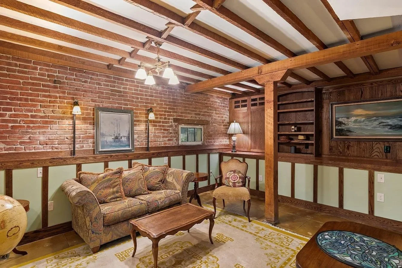 Living room with brick wall, wooden ceiling beams, beige patterned sofa, upholstered armchair, wooden coffee table, area rug, wall-mounted lamps, painting, small window, and decorative shelving.