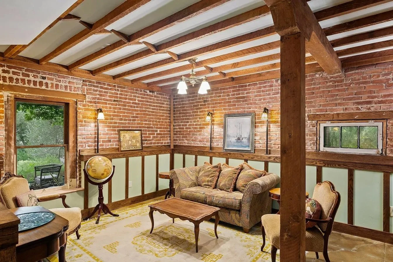 Cozy living room with brick walls, wooden beams, and vintage furniture, including a sofa, armchairs, and a globe, with windows showing greenery outside.