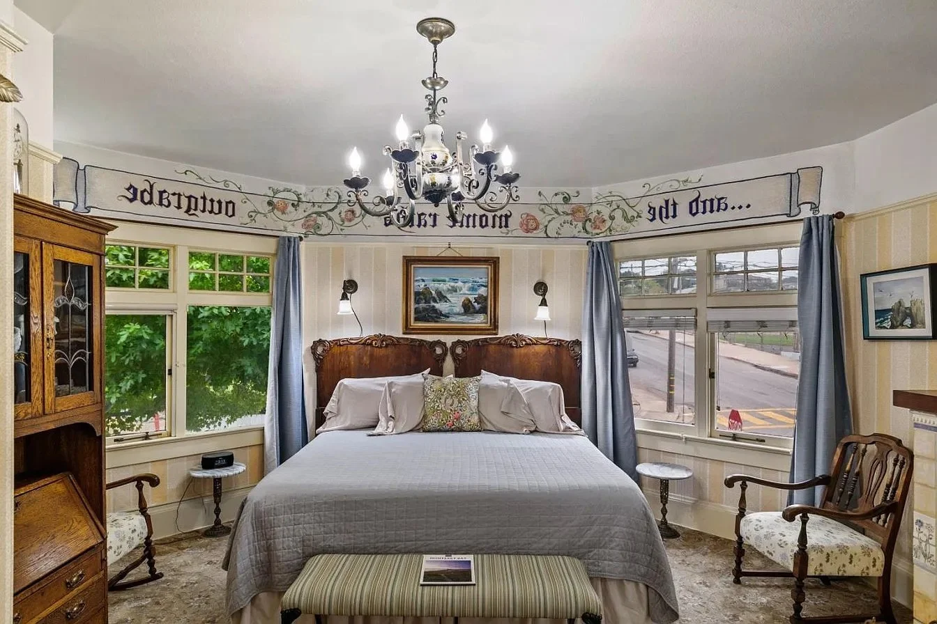 A vintage bedroom with a bed, a wooden headboard, and a chandelier. Windows with blue curtains, a framed painting of a landscape, a wooden dresser, and two armchairs with floral upholstery.