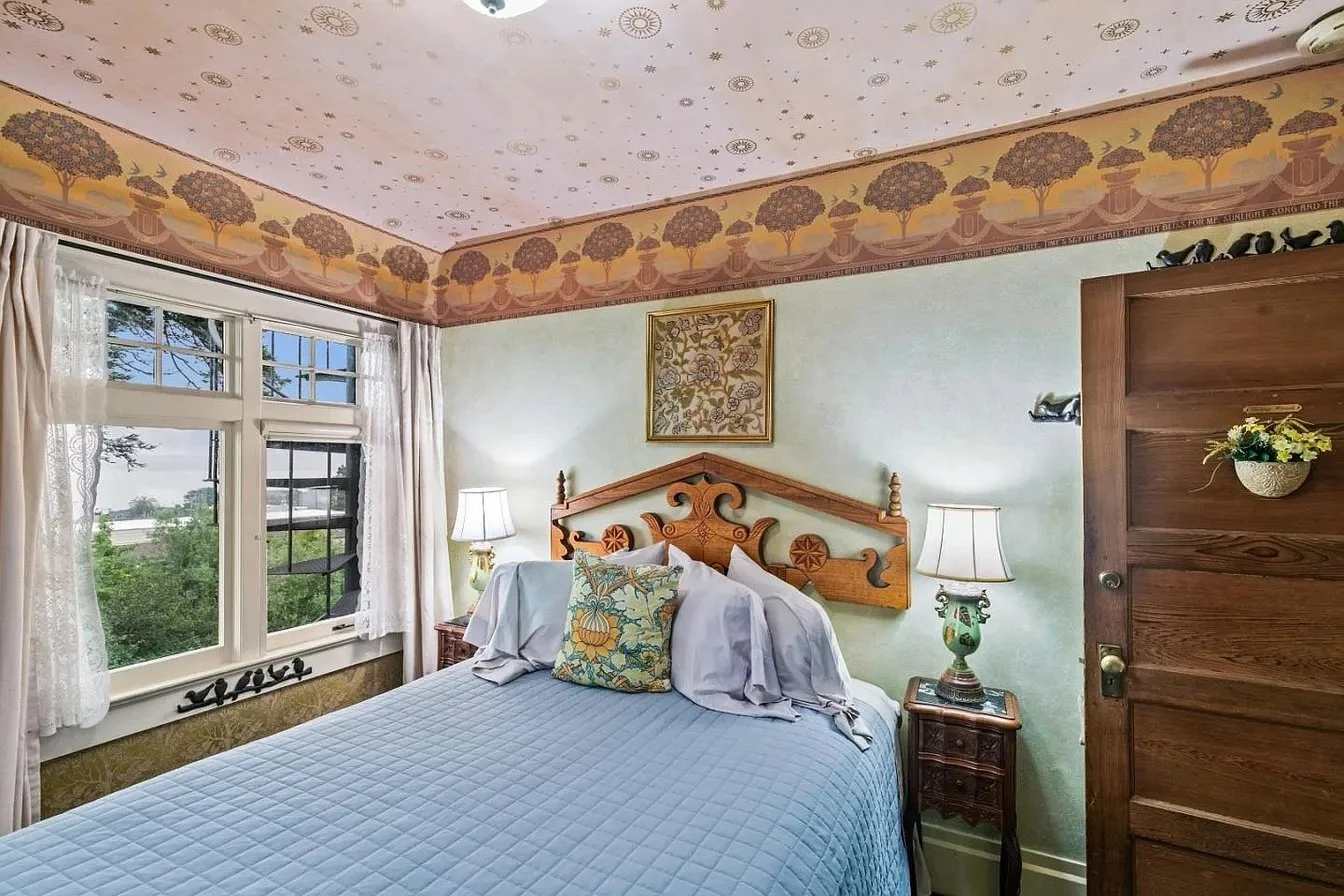 A bedroom with light green walls, a window with white curtains, a wooden headboard, nightstands with lamps, and a bed with a blue quilt and decorative pillows.