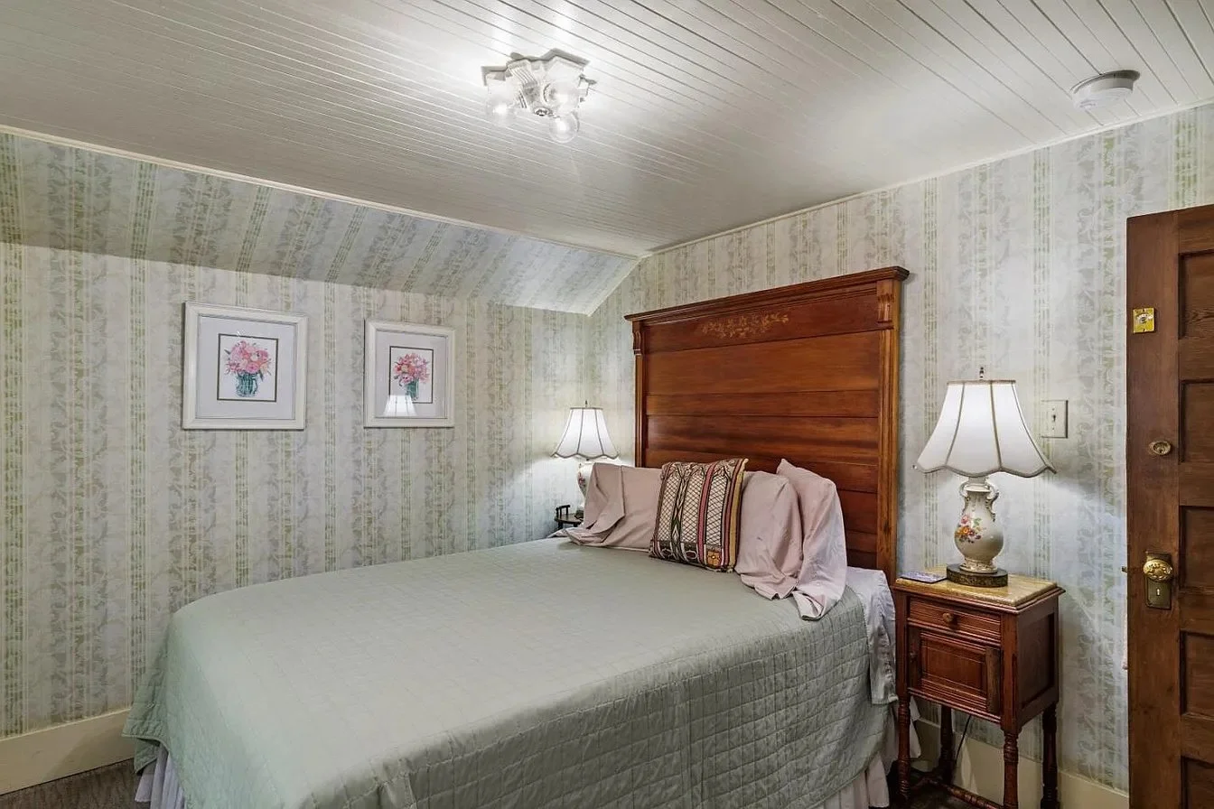 A cozy bedroom with a wooden headboard, floral wallpaper, and framed floral artwork. There are two matching lamps on nightstands on each side of the bed, and a wooden door to the right.