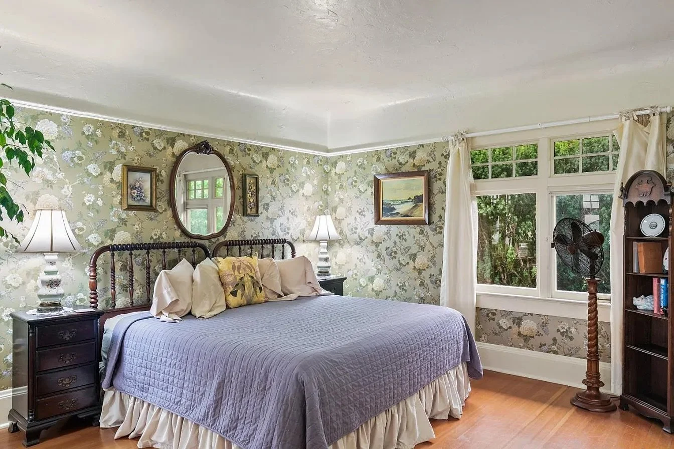 A cozy bedroom with floral wallpaper, a wooden bed with a lavender quilt, and matching pillows. Two nightstands with table lamps flank the bed. A mirror and framed artwork are on the wall, with a window allowing natural light, covered with white curtains. A fan on a wooden stand is near a bookshelf in the corner.