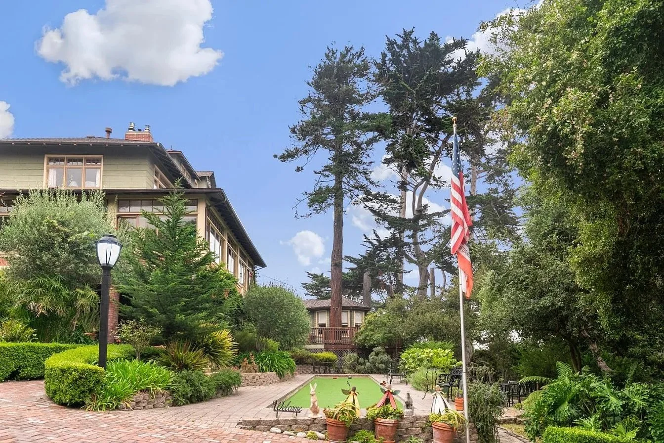 A landscaped garden with a brick walkway, small bushes and trees, and a flagpole with an American flag. There is a large tree in the background and a building with large windows on the left. Small decorative rabbits and other ornaments are near the f