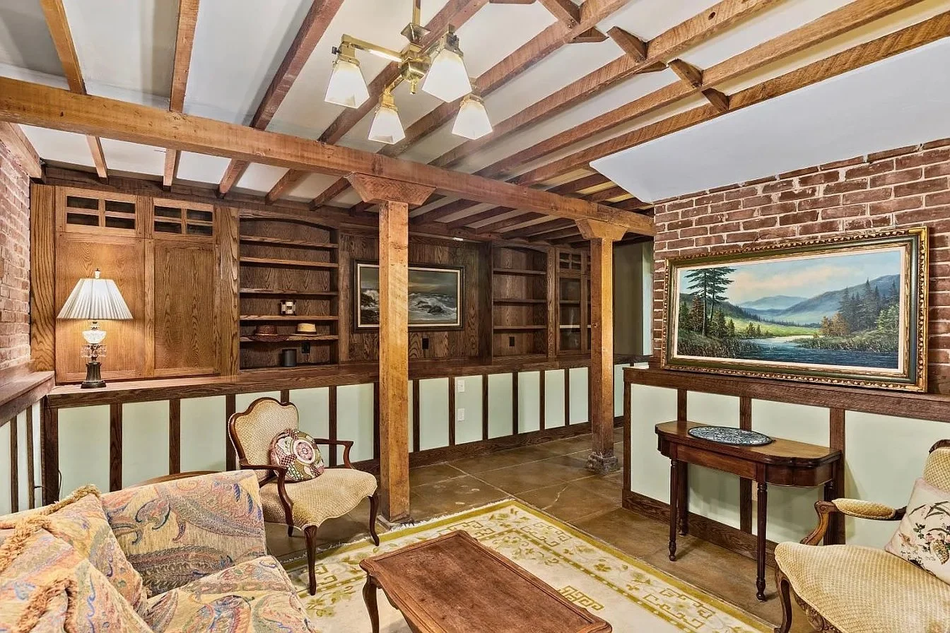 Living room with wood beams, brick walls, traditional furniture including a floral sofa, armchair, and wooden table, paintings on the walls, and a patterned rug.