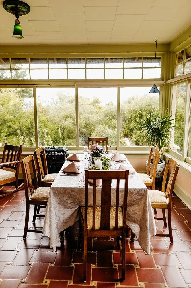 A dining room with a table set for a meal, featuring nine wooden chairs, a floral centerpiece, candles, and pink napkins. The room has large windows showing trees outside, and a green ceiling light hanging above.