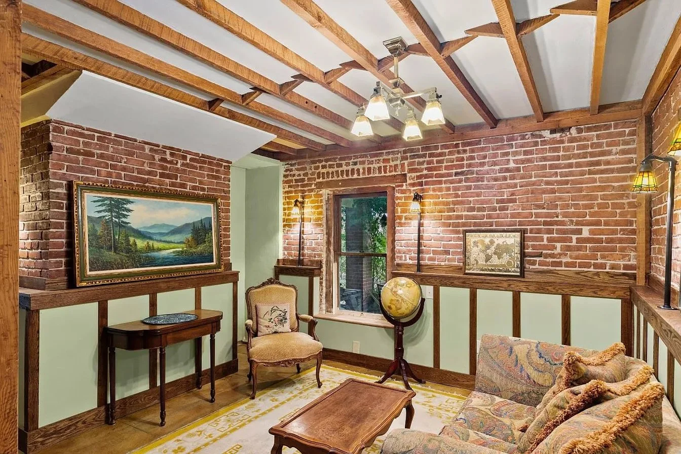 Living room with exposed brick walls, wooden ceiling beams, vintage furniture, a painting of a landscape, a globe, and brass lamps.