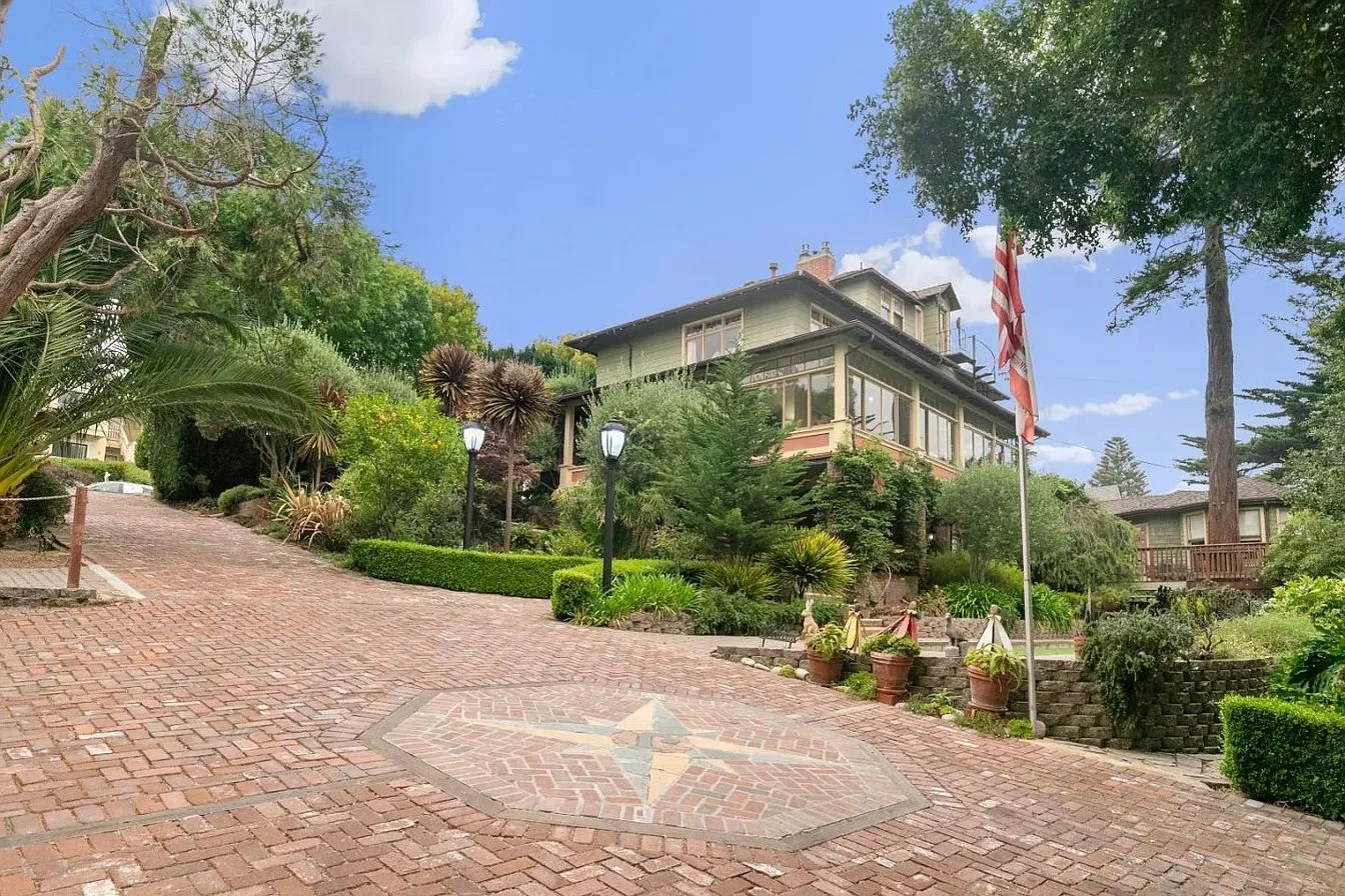 A large American-style house on a hillside surrounded by greenery, with a brick driveway, lamp posts, potted plants, and an American flag flying.