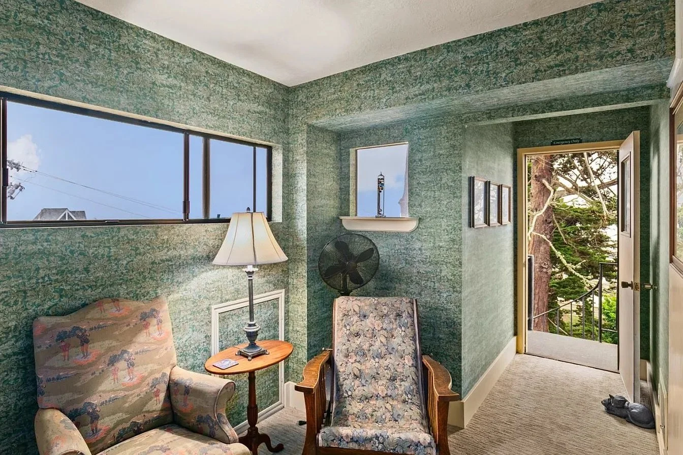 Room with green textured walls, large window, floral upholstered armchair, floor lamp, small wooden table, vintage chair, black fan, open door leading outside to trees, and a pair of shoes on the floor.