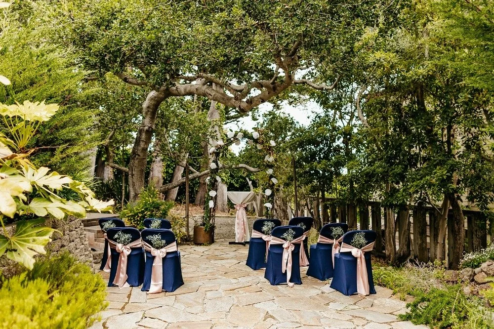 Outdoor wedding setup with six chairs decorated with pink ribbons and small white flowers, arranged around a stone-paved area under a large tree with lush green foliage, and a decorated arch at the center with hanging white flowers.