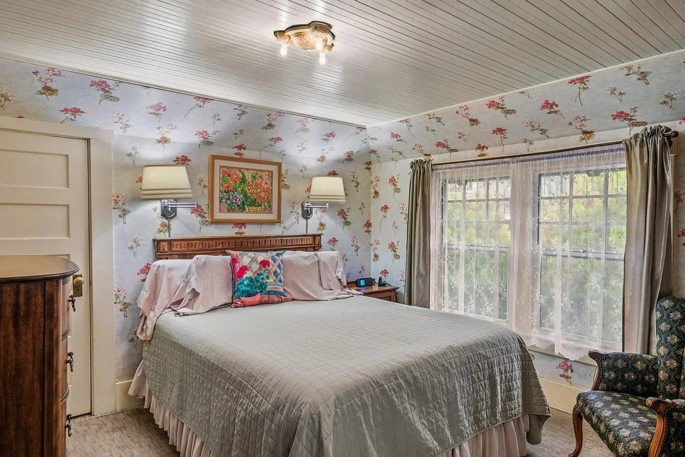 A bedroom with floral wallpaper, a bed with a quilted bedspread, pillows, and a wooden headboard. There are two wall-mounted lights, a painting above the bed, a nightstand with a clock and phone, a curved armchair with floral upholstery, and a large window with lace curtains and green drapes.