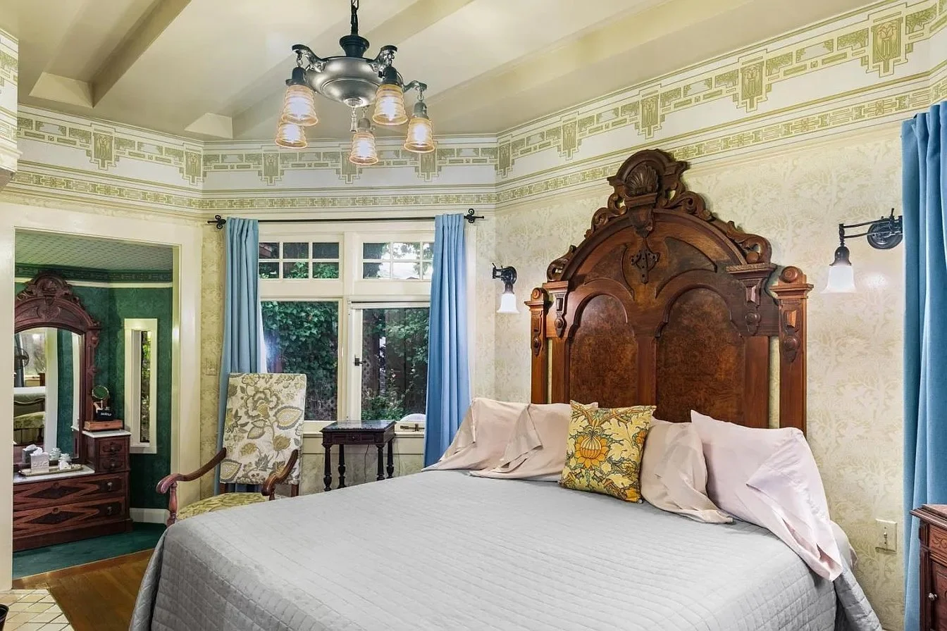 A vintage bedroom with a large bed, ornate wooden headboard, blue curtains, a floral armchair, a small wooden nightstand, a dresser with mirror, and a window showing greenery outside.