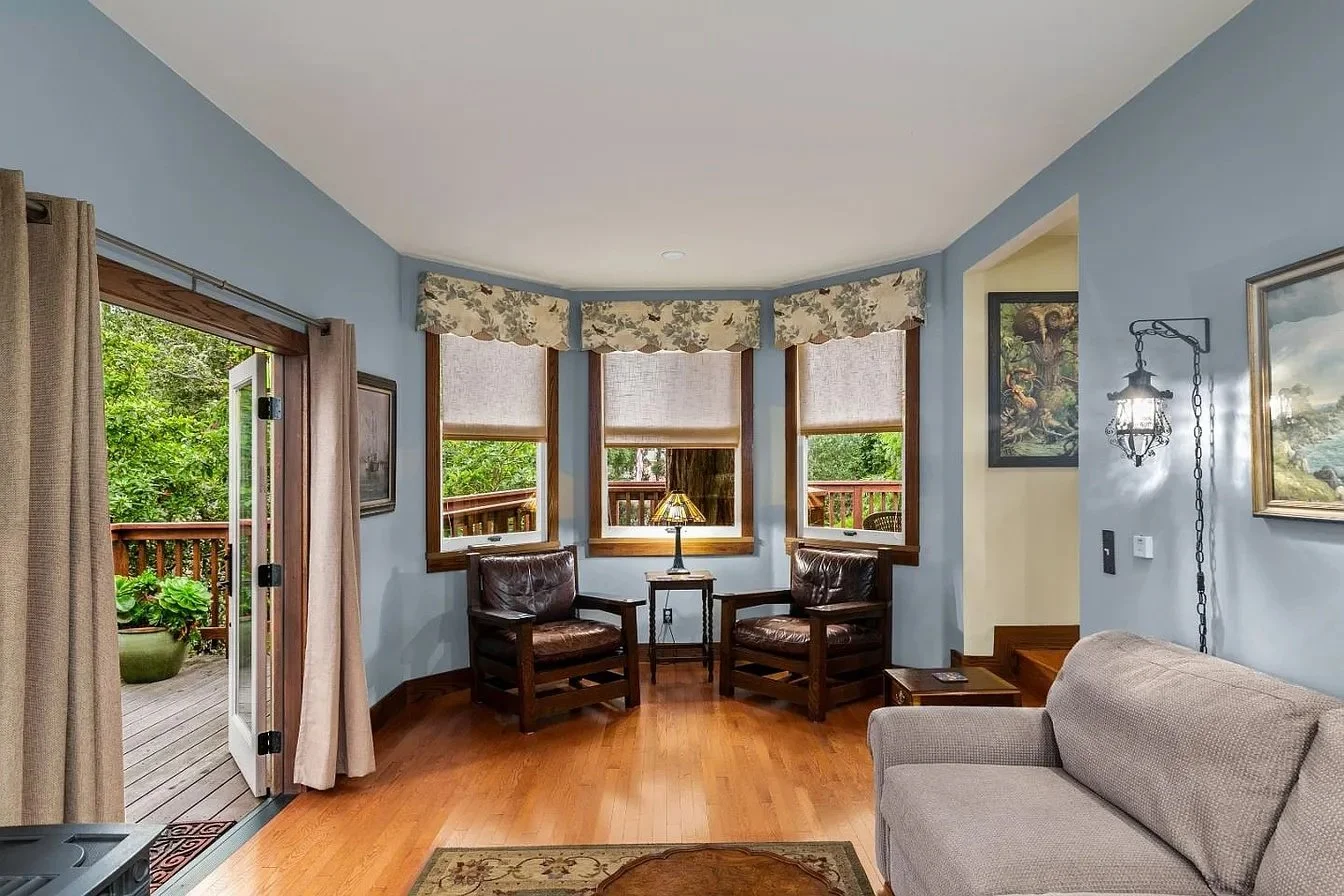 A cozy living room with light blue painted walls, hardwood flooring, and a sliding glass door leading to a wooden deck with plants. The room has a beige sofa, a small wooden side table, two dark wood chairs with leather cushions, a table lamp with a stained glass shade, and several wall-mounted paintings. There is a large bay window with floral valances and blinds.