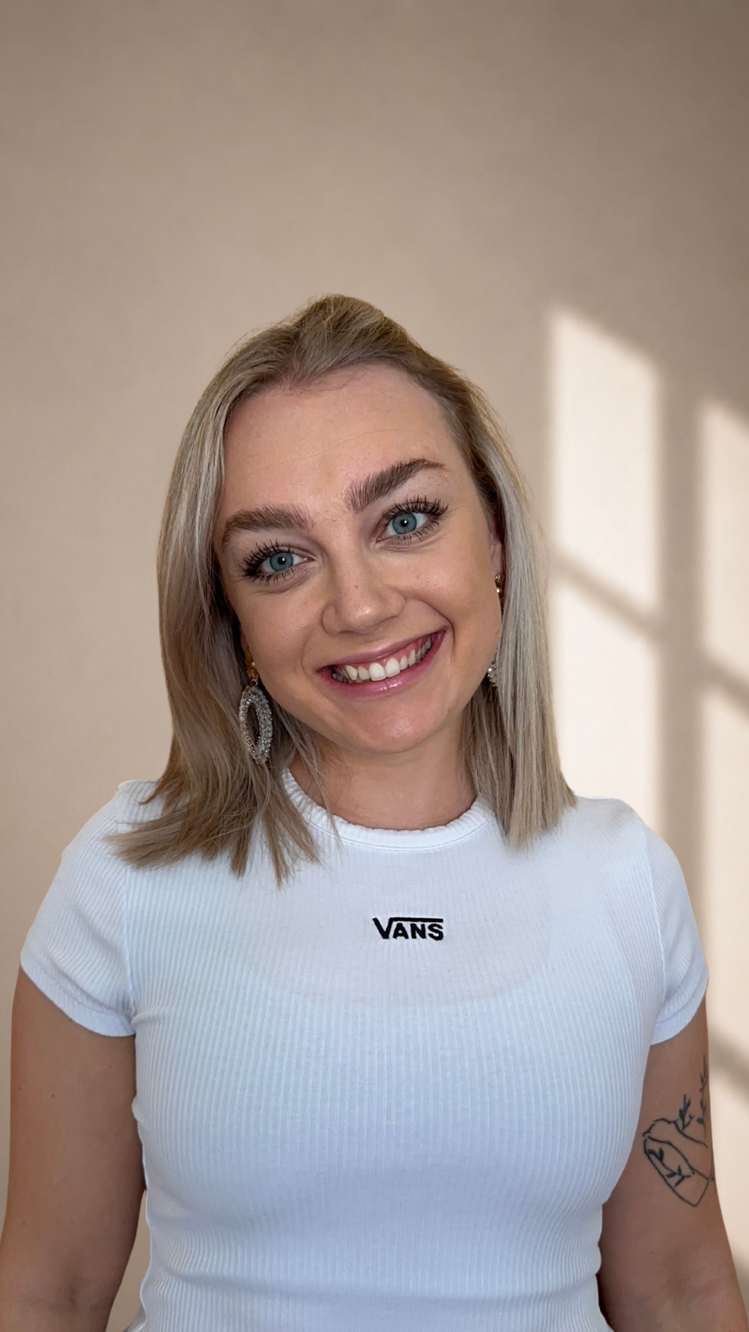 Eine junge Frau mit blonden Haaren, blauen Augen und Ohrringen lächelt vor einer beigefarbenen Wand, auf die das Sonnenlicht sanfte Schattenmuster wirft. Sie trägt ein weißes T-Shirt.