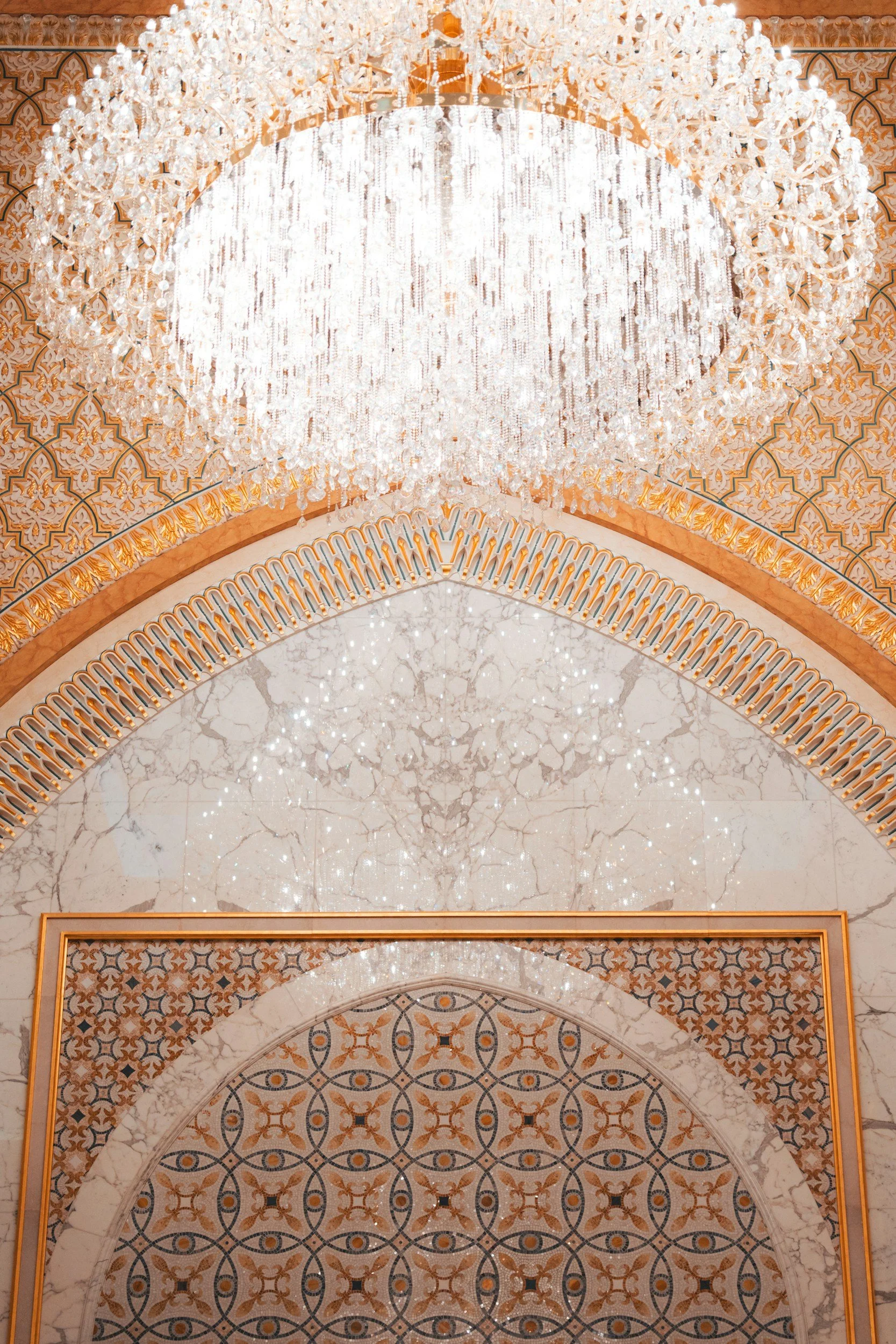 A luxurious chandelier hangs from a ornate ceiling featuring intricate patterns in gold, black, and white, with marble flooring below.