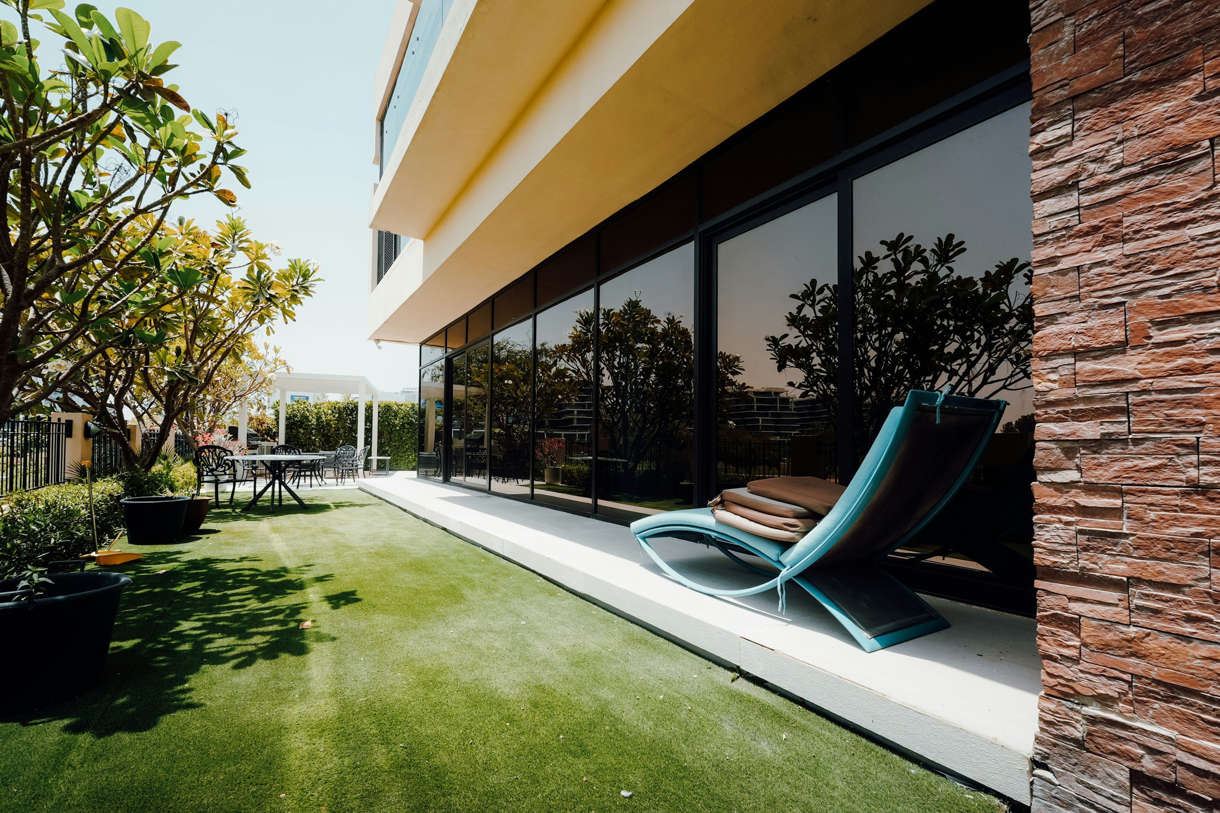 Residential patio area with outdoor lounge chair, glass facade reflecting trees, and landscaped yard with small trees and patio furniture.