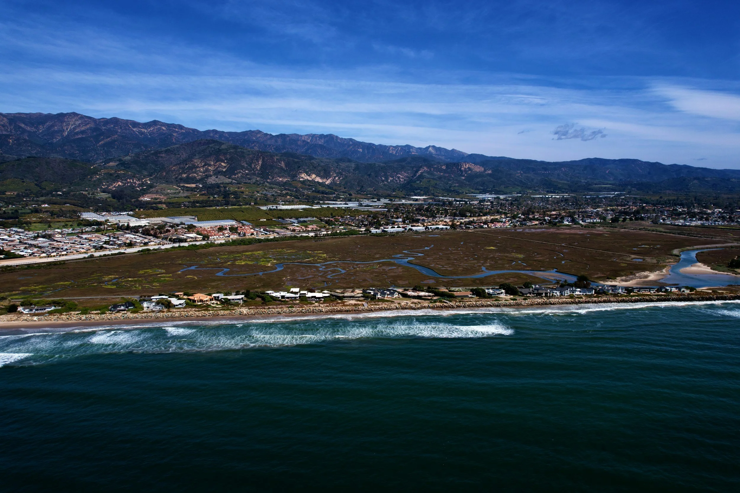 Scenic view of a coastline with waves hitting the shore, marshland with ponds and roads, residential houses, and mountains in the background under a partly cloudy sky.