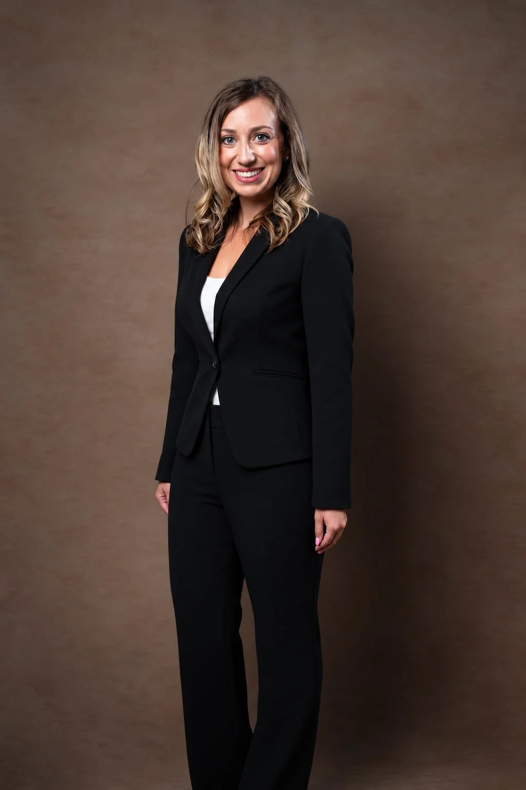 A woman in a black business suit smiling, standing against a brown background.