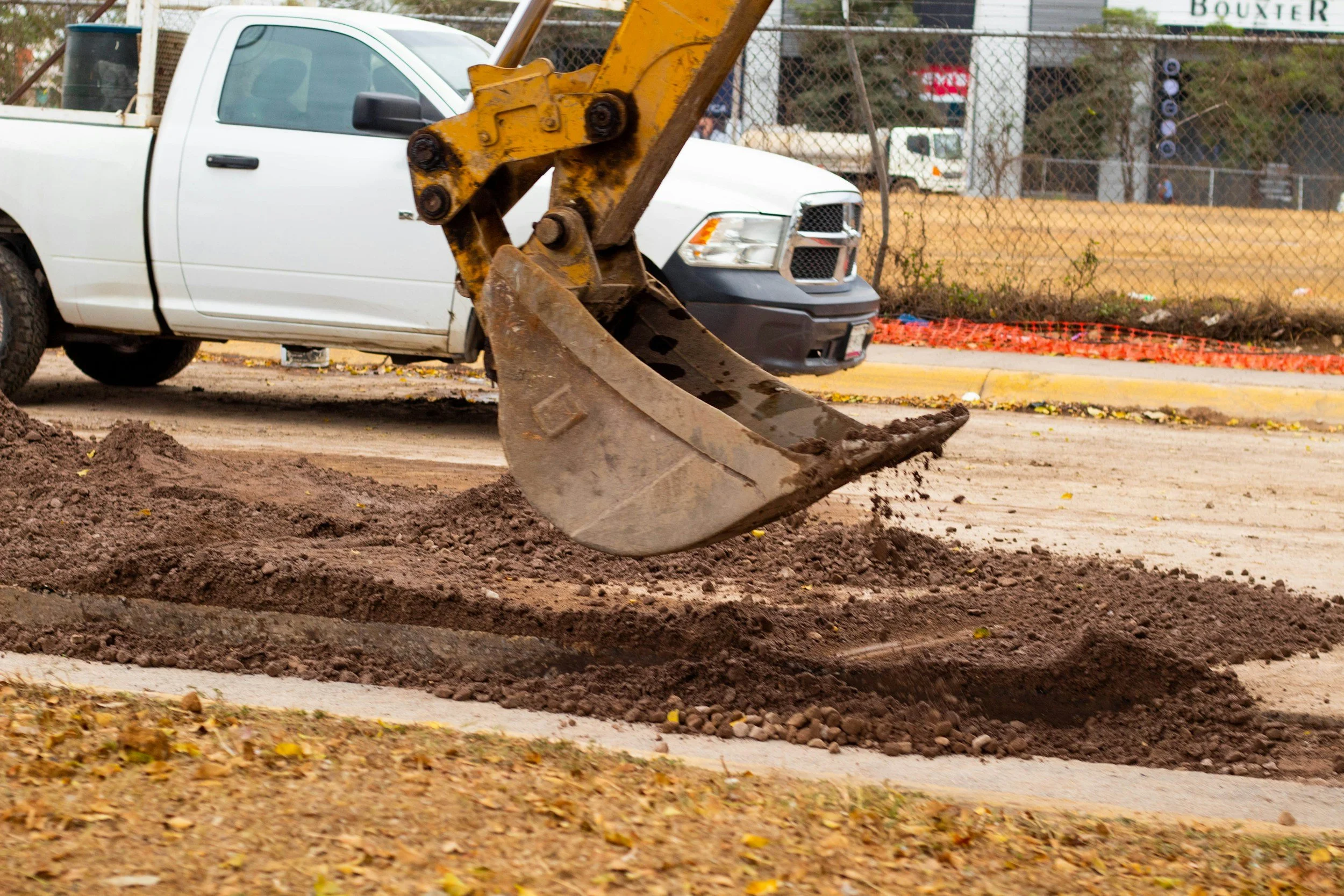 Construction site with a white pickup truck and a yellow excavator digging into the ground, surrounded by a chain-link fence and orange safety fencing.