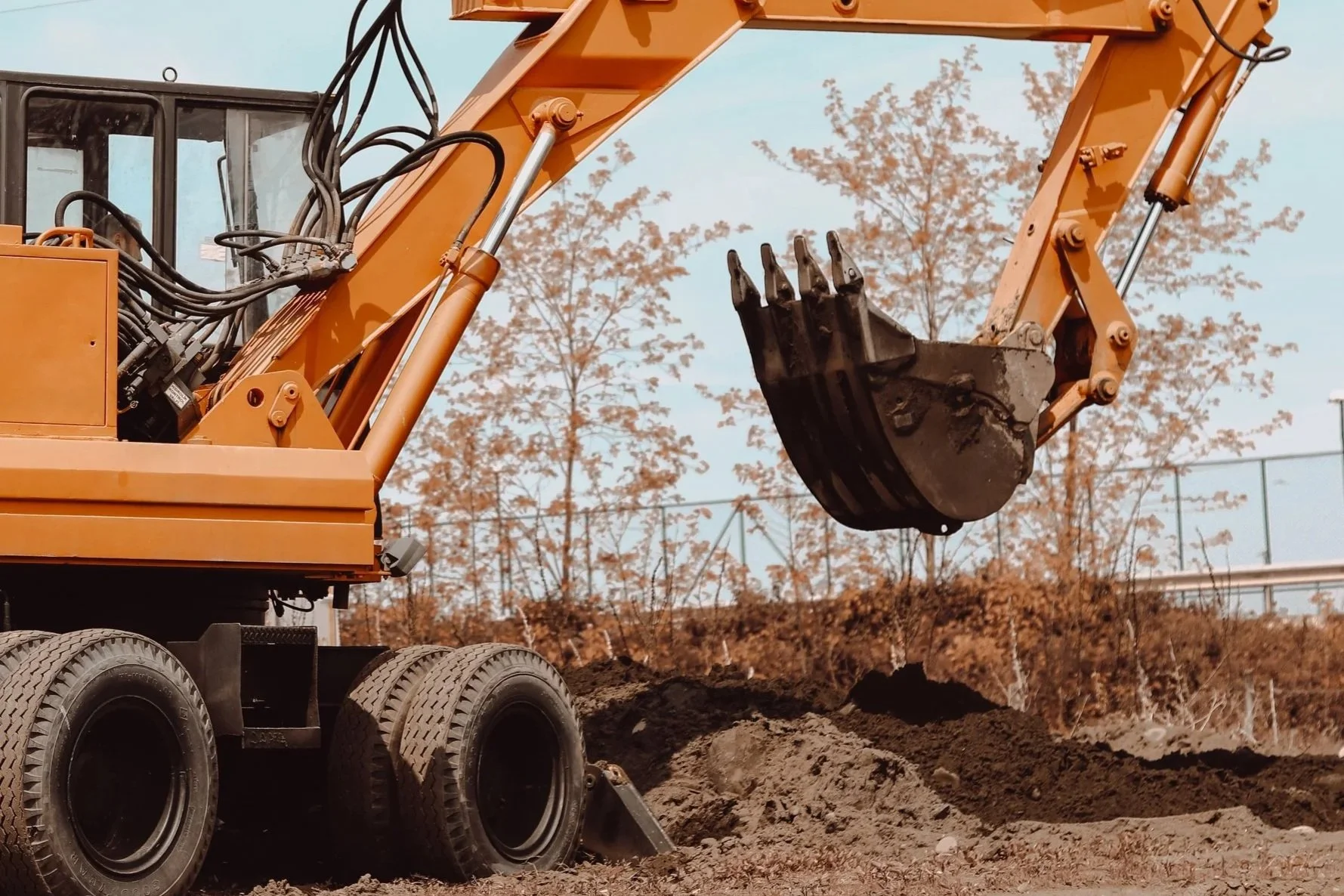 Construction excavator with a bucket, moving dirt at a construction site with trees and a fence in the background.