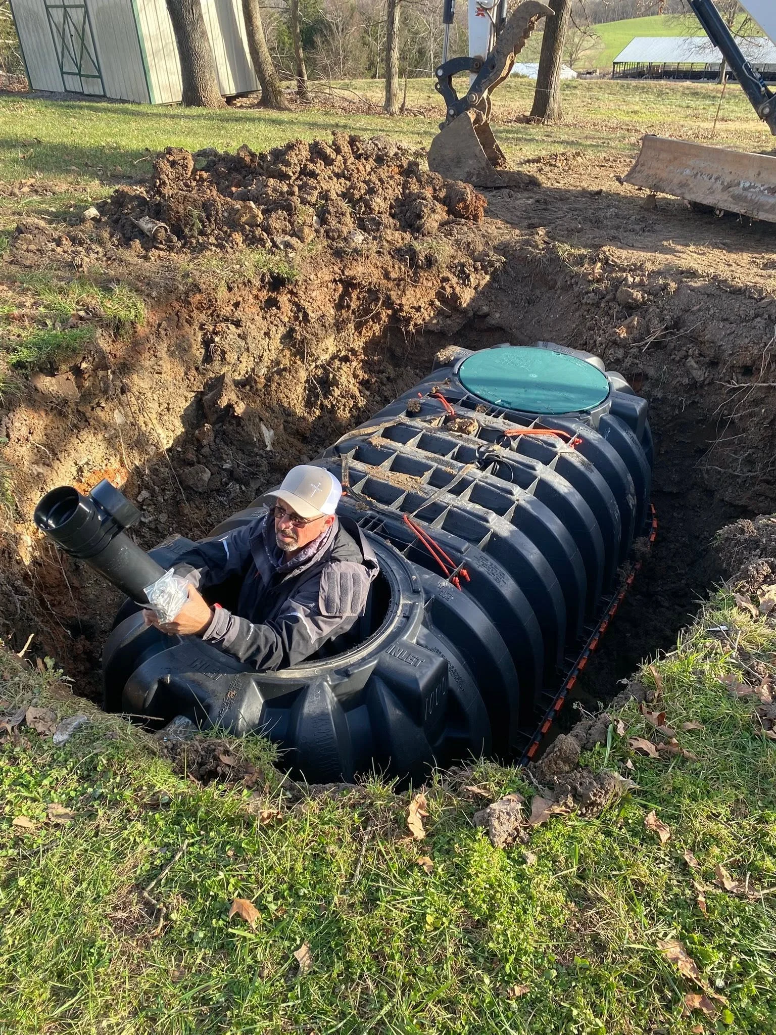 A man working outdoors next to a large black septic tank, digging a hole in the ground with a backhoe. There are trees, a grassy area, and a shed in the background.