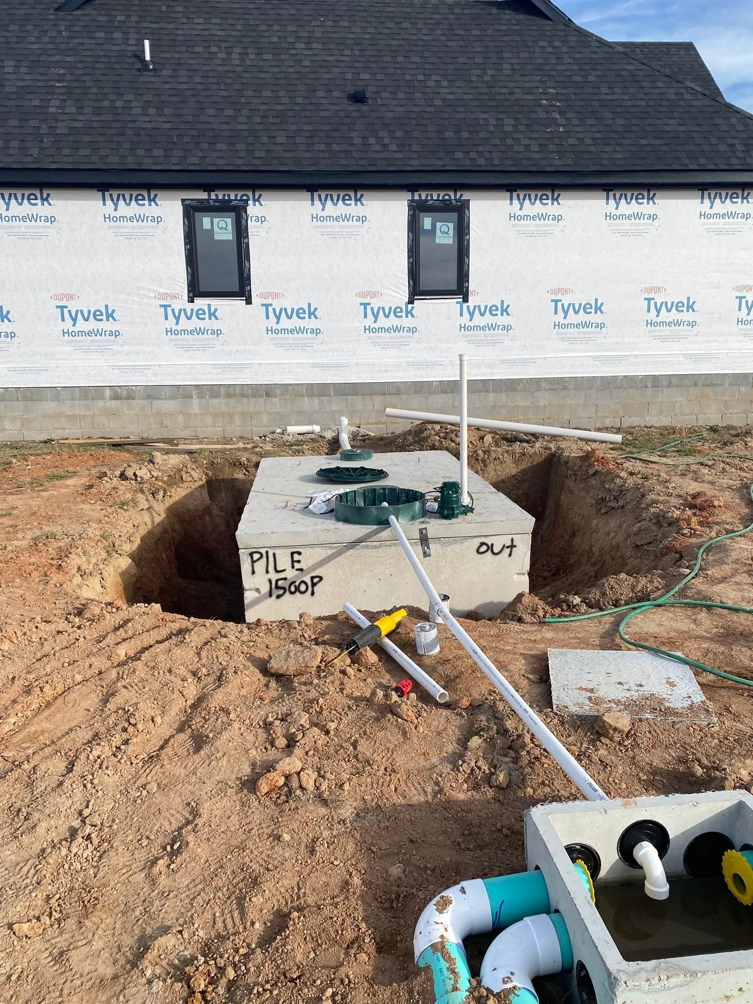 Underground plumbing work in progress at a construction site, with exposed PVC pipes and a sump pump, in front of a house under construction.