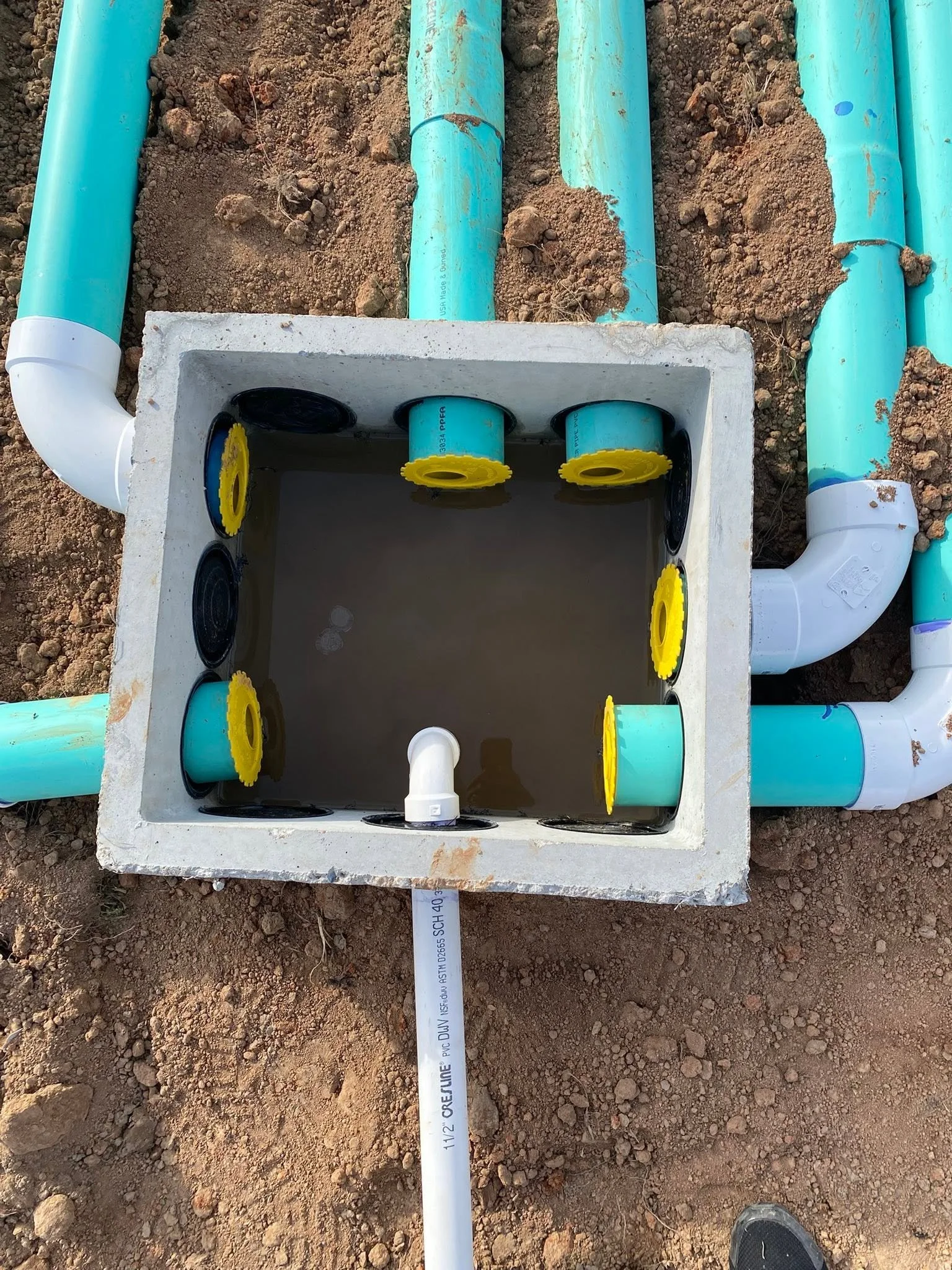 The image shows a construction site with a trench and PVC pipes laying in dirt. The pipes are connected to a concrete box with black and yellow fittings and some water inside the box. There is a white PVC pipe extending out of the box with some text on it.