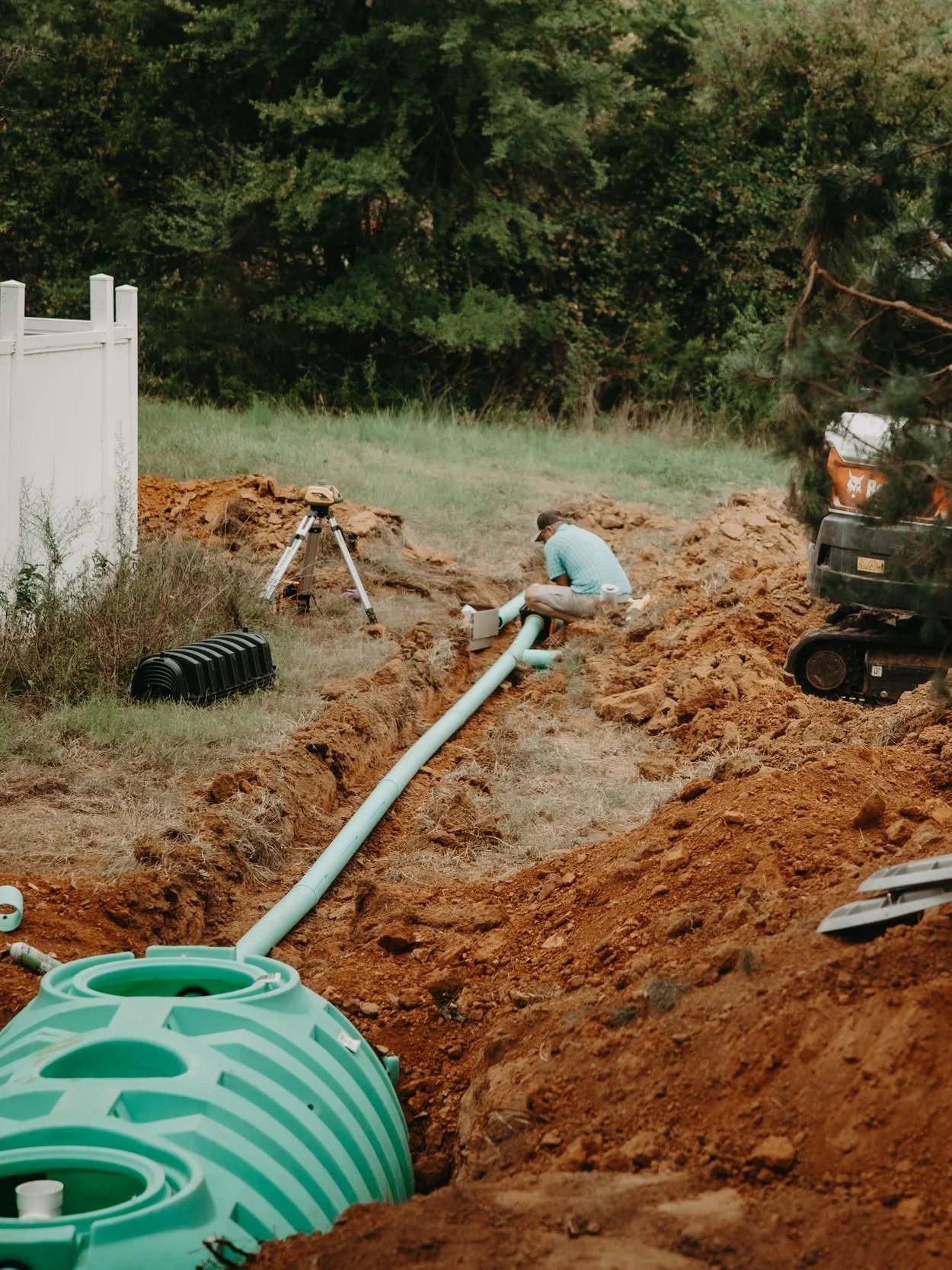 A person working on a plumbing installation or repair in an outdoor area, with construction equipment, pipes, and a trench in the ground.