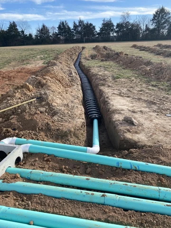 A construction site showing a trench with black and blue pipes being installed in the ground, with dirt piled along the trench and a grassy field with trees in the background.