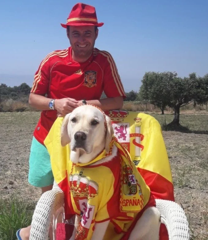 A man wearing a red Spain soccer jersey and a red hat is smiling, standing behind a dog draped in a Spanish flag, sitting on a chair outdoors on a sunny day.