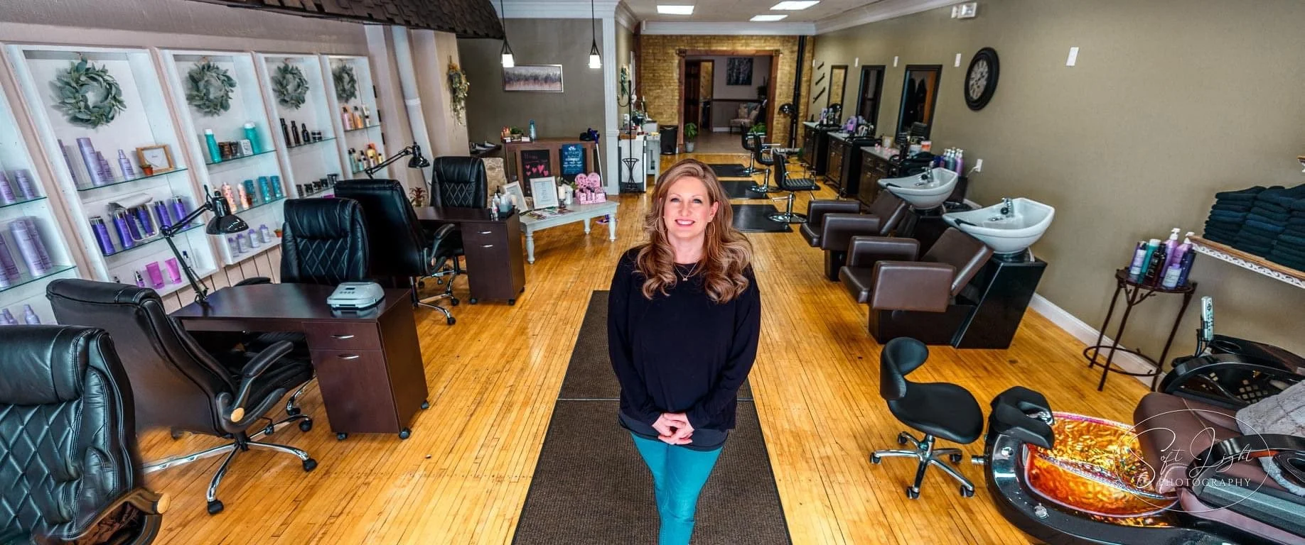 Interior of a hair salon with hair washing stations, shelves with hair products, and styling chairs. A woman with long, wavy hair and a black top is smiling at the camera in the center of the room.