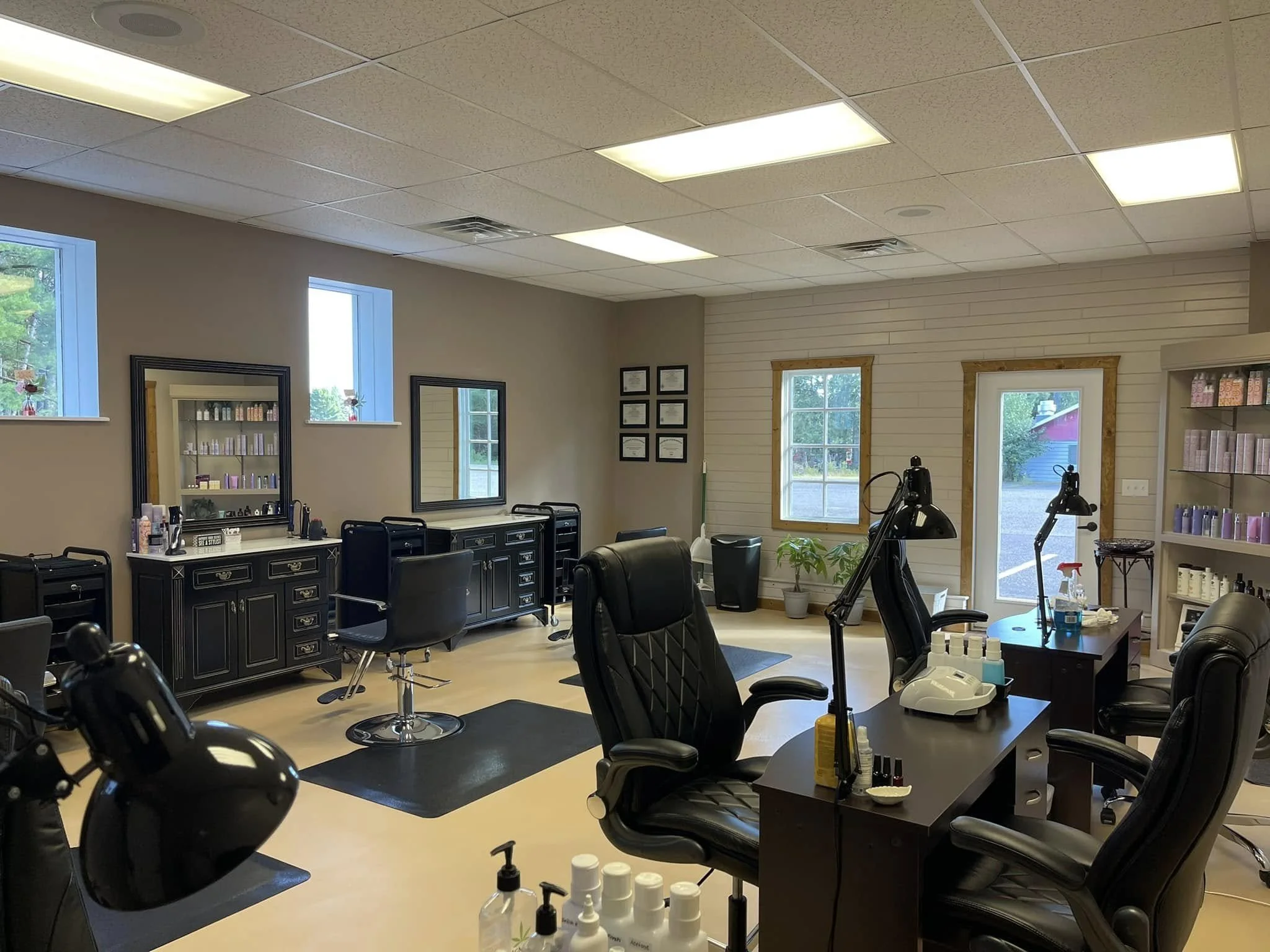 Interior of a beauty or wellness salon with multiple massage chairs, black cabinets, and shelves with products. There are windows letting in natural light and framed certificates on the wall.