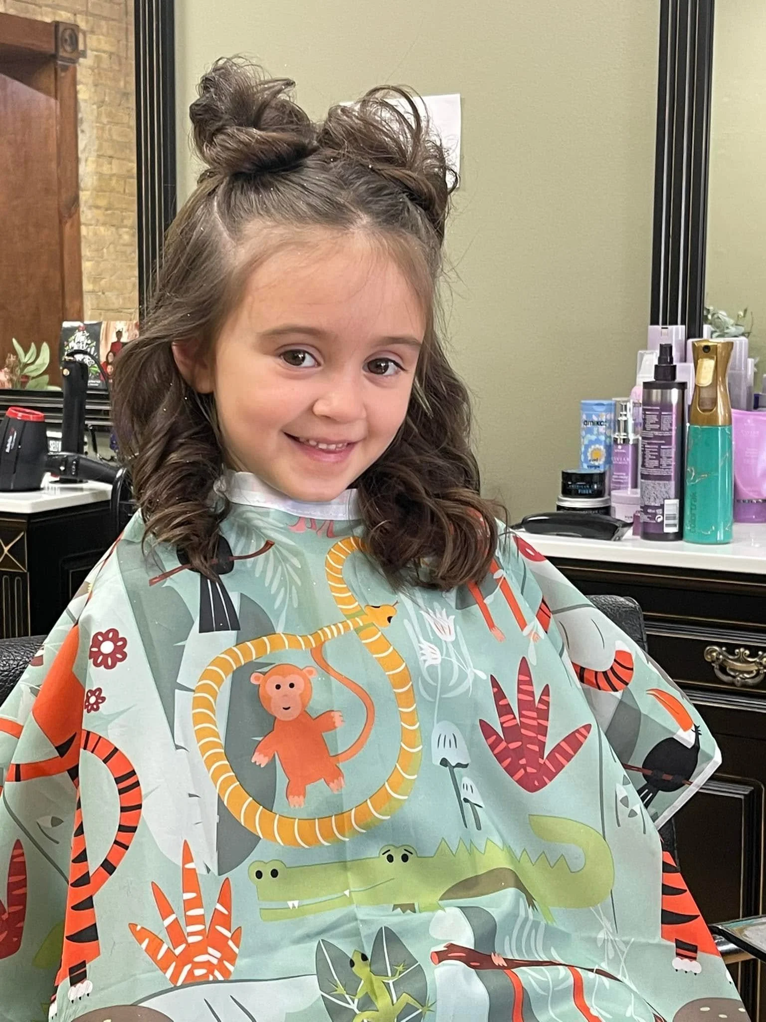 Young girl with brown curly hair sitting in a salon chair, smiling, wearing a colorful cape with jungle animals and plants design. Cuts and styles at FABCO salon. Fallon Aliece Beauty Co. Eagle River, WI 