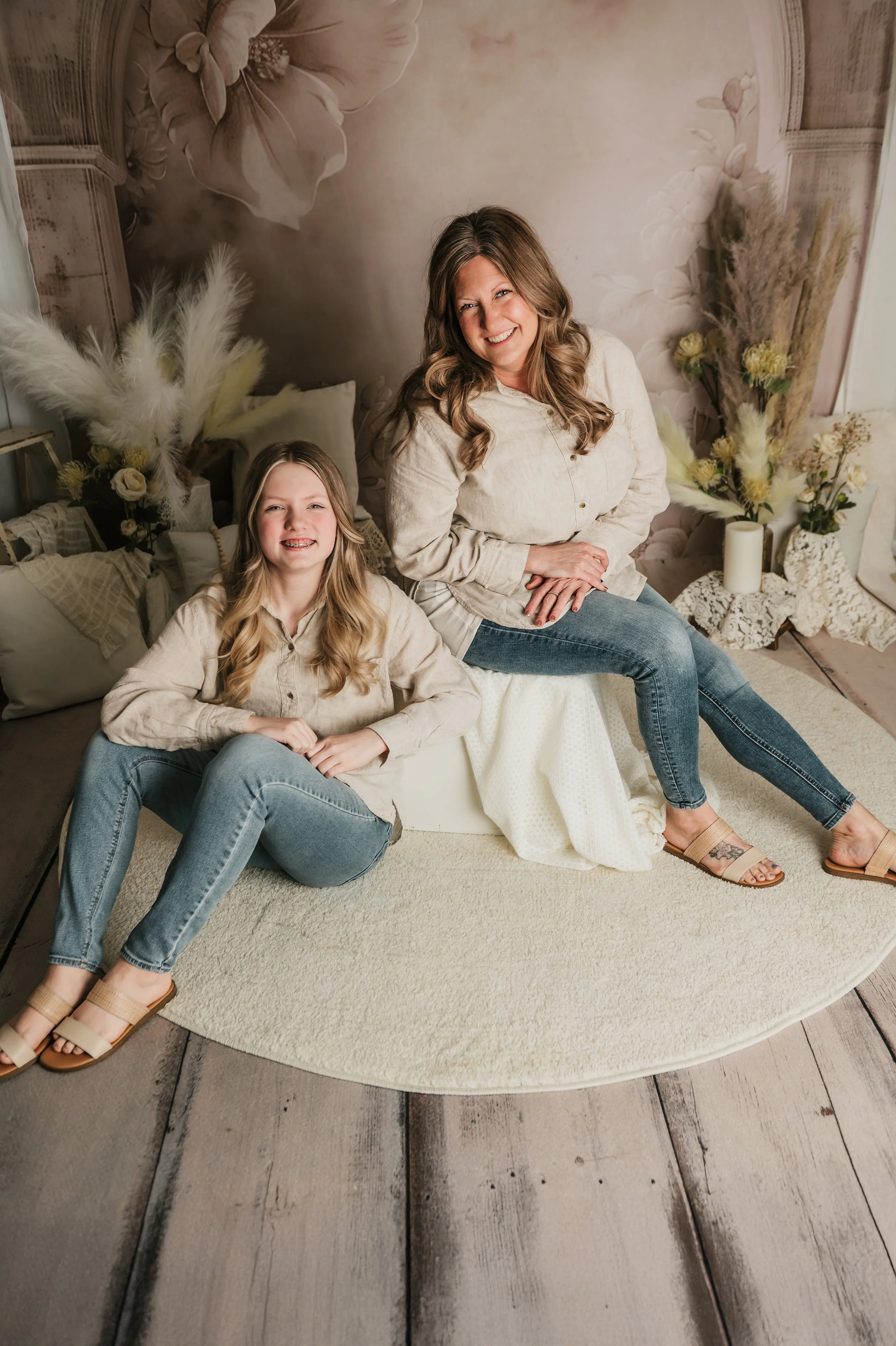 A woman and a girl sitting on a round beige rug on a wooden floor, smiling at the camera, with a floral-themed wall behind them, decorated with vases of dried flowers and candles.