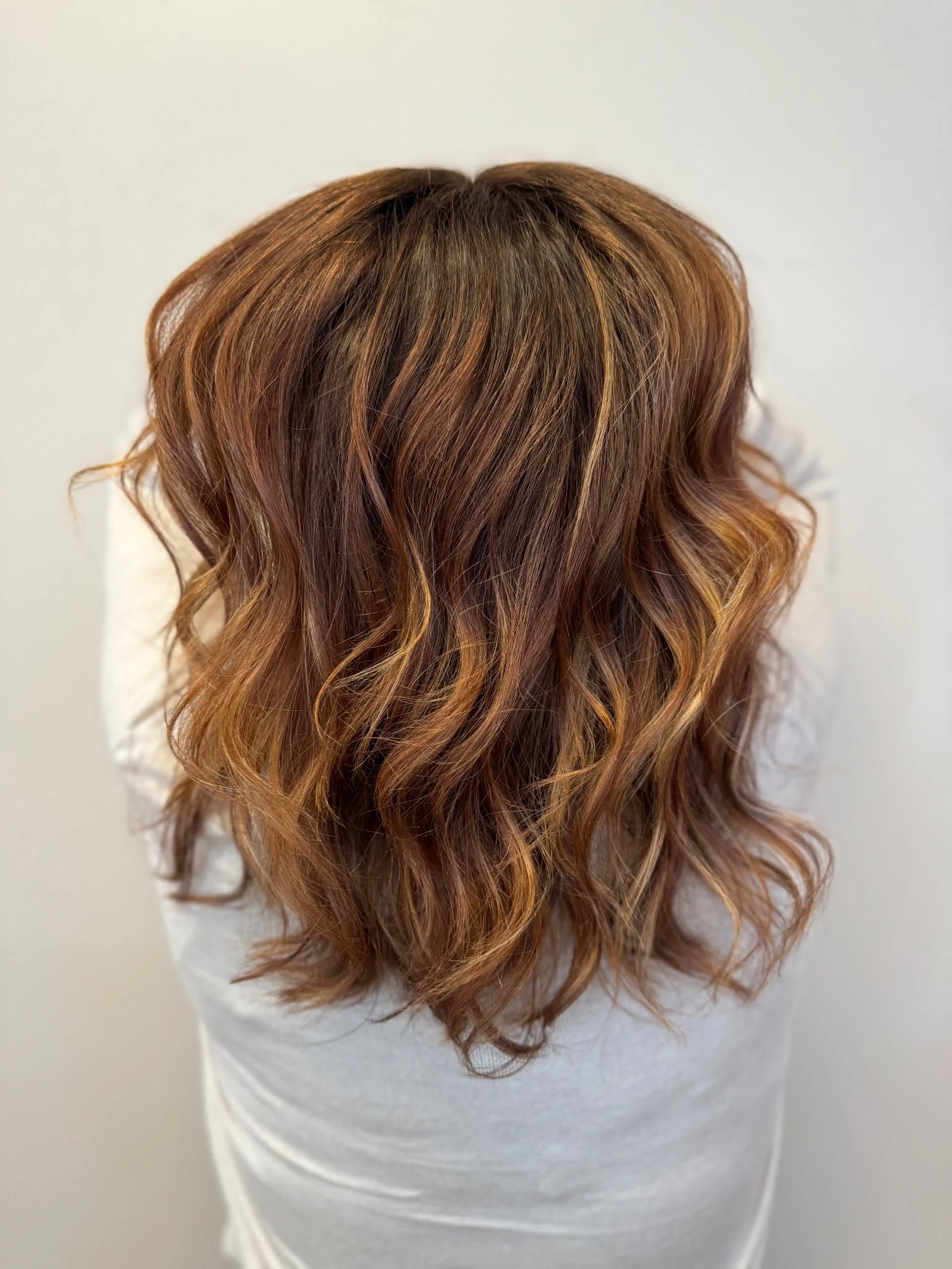 Top view of a person with long, wavy, brown hair with lighter highlights, wearing a white shirt. Color and styling in Eagle River, WI. Fallon Aliece Beauty Salon