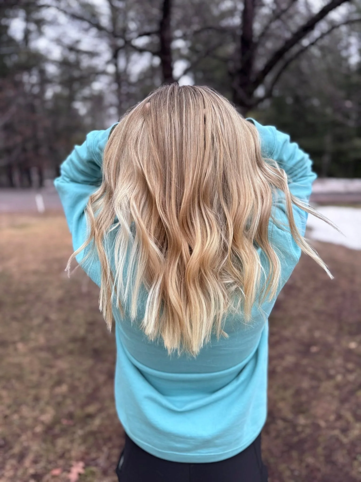 A person with long blonde hair wearing a blue hoodie, standing outdoors with their back to the camera and their hands behind their head. The background features trees and a cloudy sky. Blonding, coloring, and styling in Eagle River, WI. 