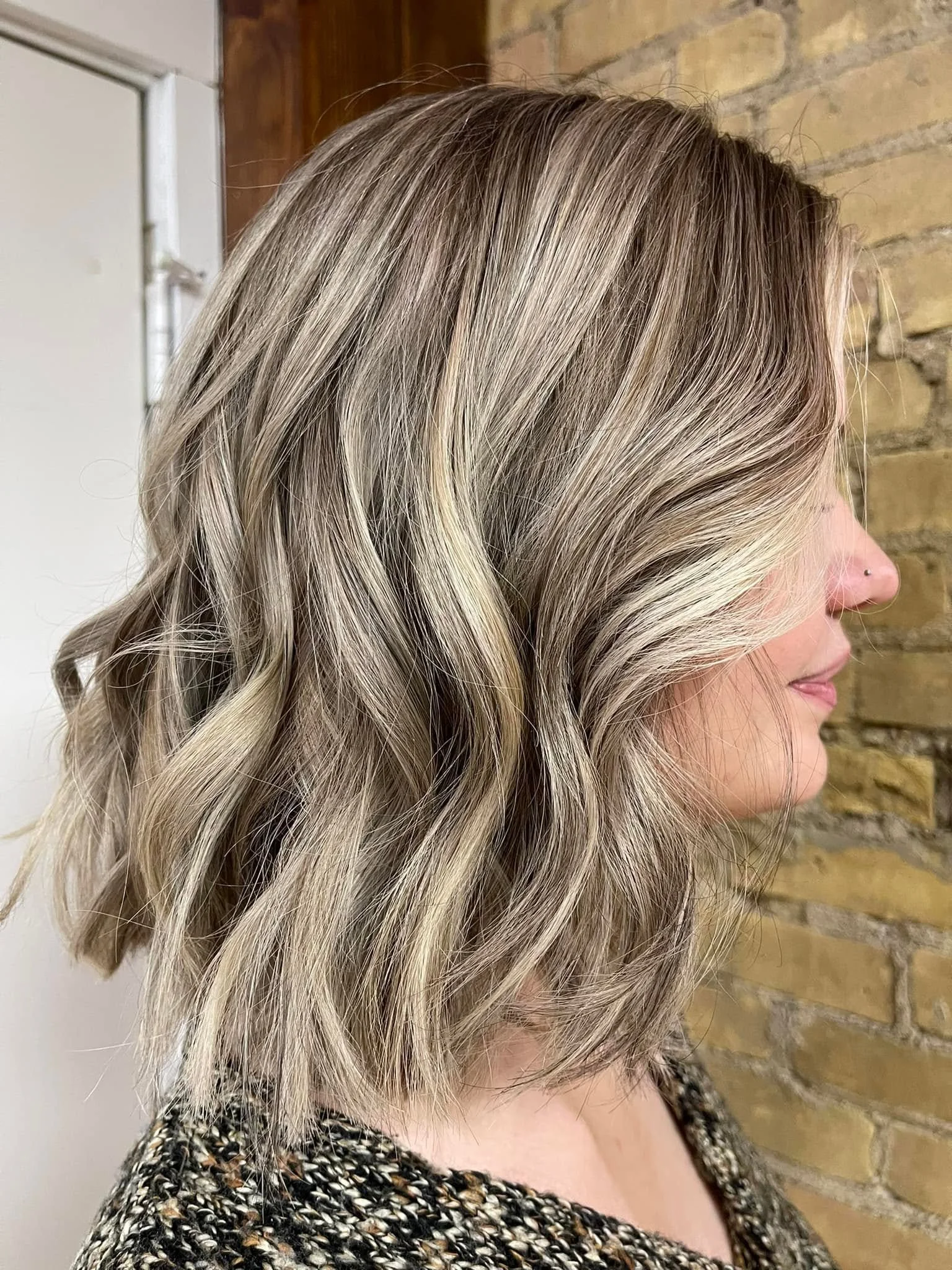Side profile of a woman with shoulder-length, wavy blonde hair in front of a brick wall. Blonding stylist. Eagle River, WI. FABCO salon