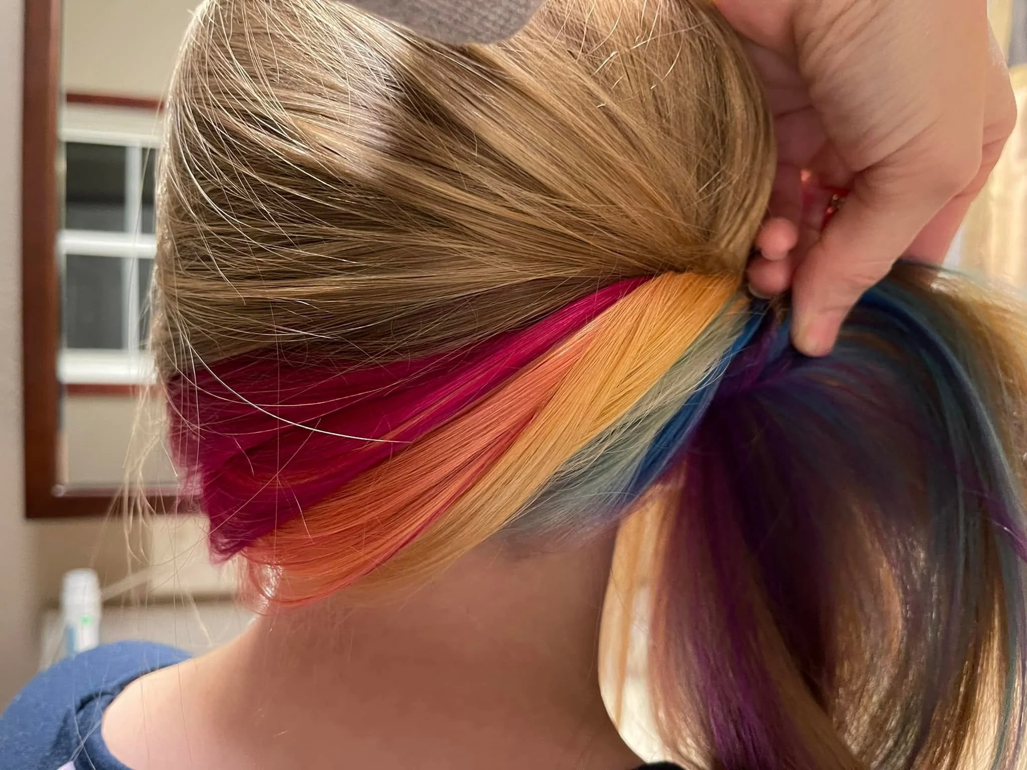 A person with multicolored hair, including shades of blonde, orange, red, pink, purple, and blue, is shown in a close-up side profile view, with hand touching their head. Falon Aliece Beauty Co. Eagle River, WI. 
