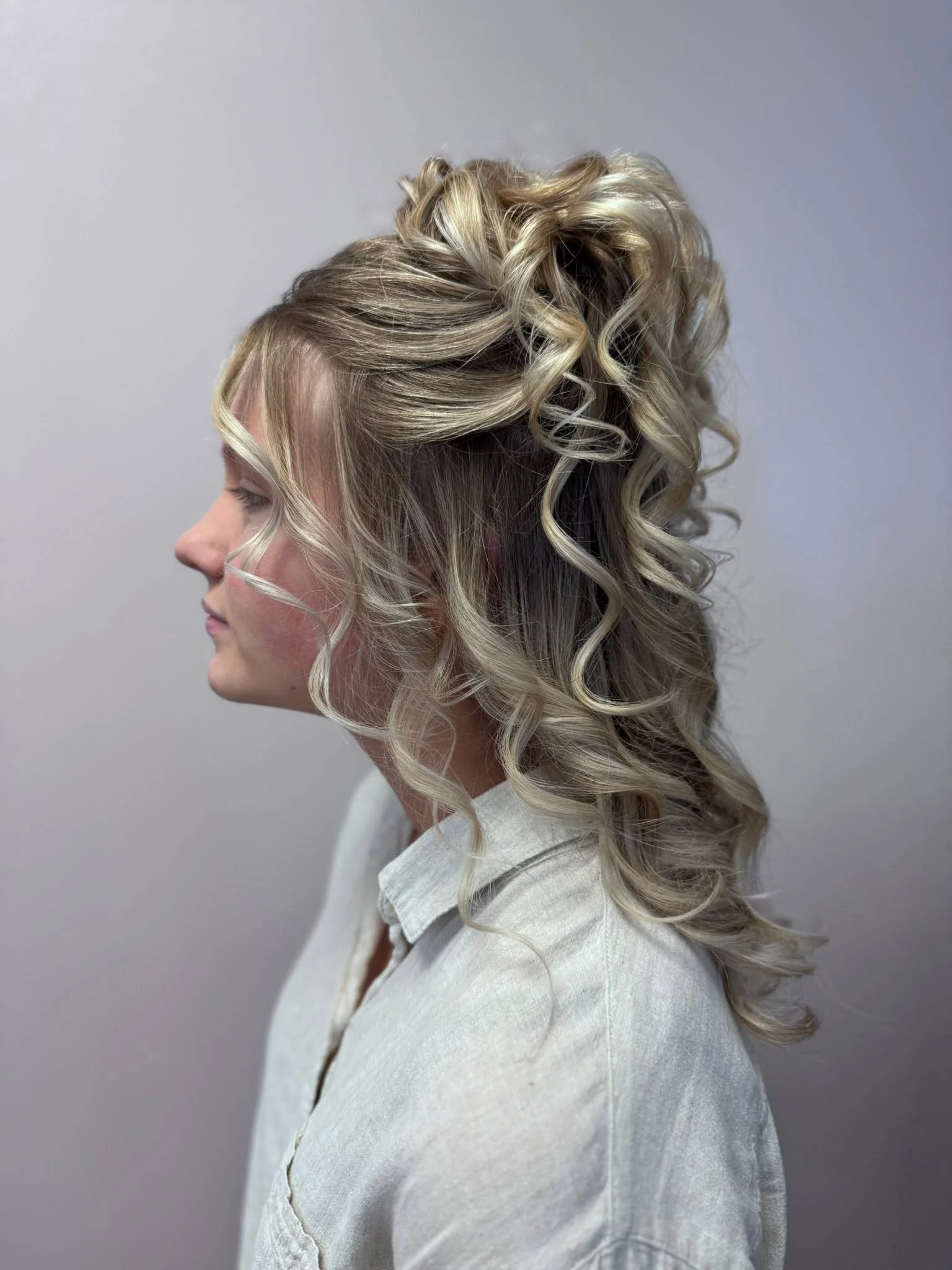 A woman with platinum blonde, curly hair and side-profile pose wearing a white blazer against a plain gray background. Updos in Eagle River, WI. Fallon Aliece Beauty Co. 