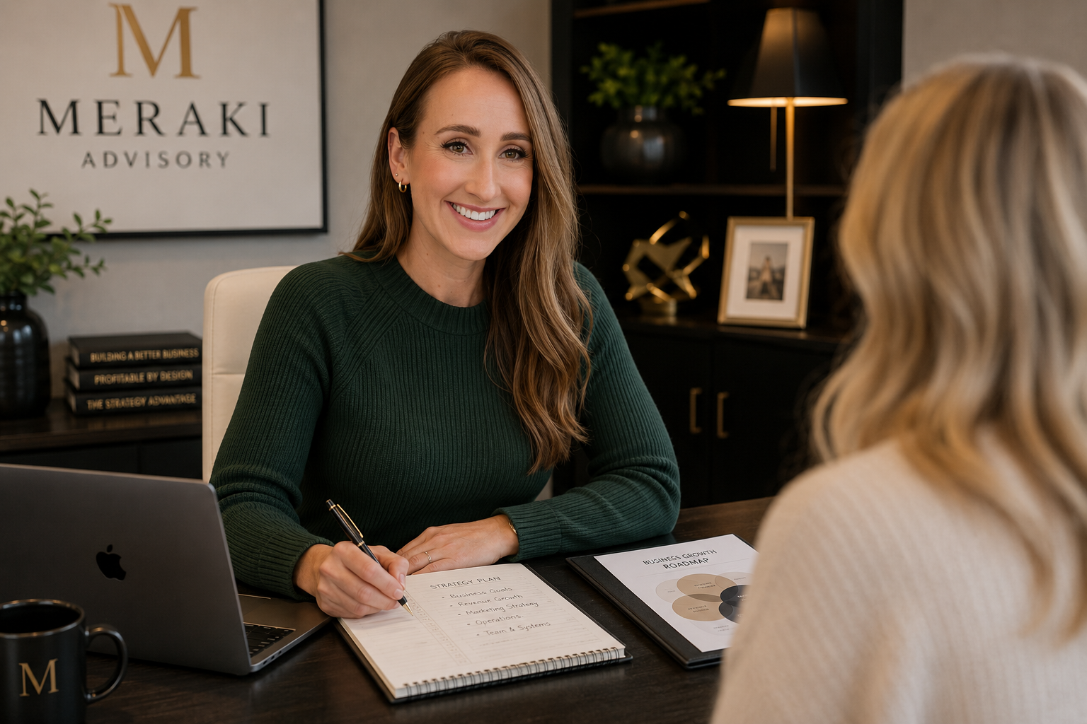 A woman with long brown hair, wearing a green sweater, is sitting at a desk, smiling, holding a pen, with a laptop and notes in front of her. She is speaking with another woman whose back is to the camera. The background features a wall with a framed logo that reads "MERAKI ADVISORY," and shelves with decorative items including books, a lamp, a framed photo, and plants.