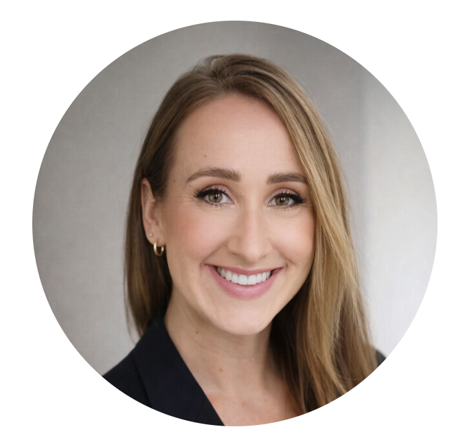 A woman with light brown hair, fair skin, and makeup, smiling in front of a plain gray background, wearing a black blazer and small hoop earrings.