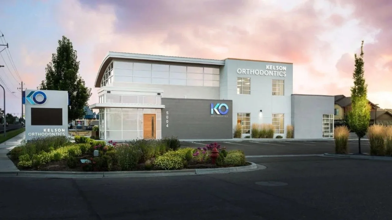 A modern orthodontics clinic named Kelson Orthodontics with a sign displaying the KO logo, located on a landscaped property with plants and flowers, under a pink and purple sunset sky.
