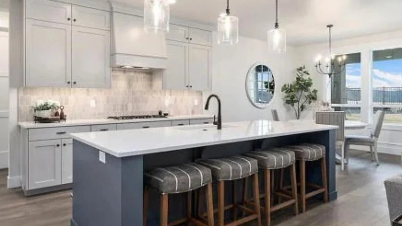 Modern kitchen with a white island, gray cabinets, beige backsplash, and round pendant lights. There is a grey-colored barstool at the island and a dining area with large windows and a view outside.