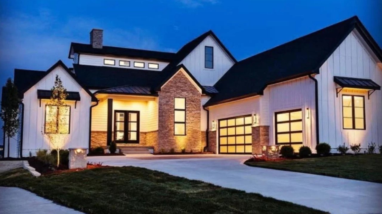 Modern house with white siding, brick accents, large windows, driveway, lawn, and outdoor lighting at dusk.