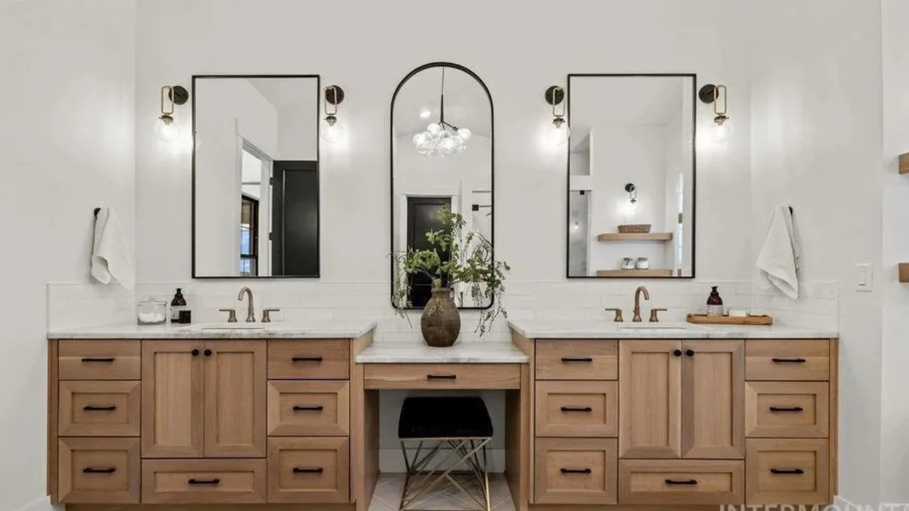Double bathroom vanity with two large mirrors, a decorative mirror in the center, and a vase with greenery between the mirrors. Light fixtures above each mirror and a stool beneath the center counter.