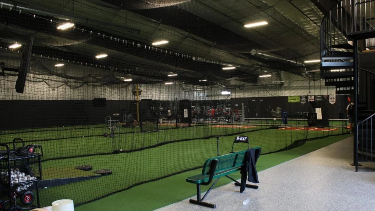 Indoor baseball training facility with artificial turf, cages, batting nets, and training equipment, including a bench and a towel hanging on it.