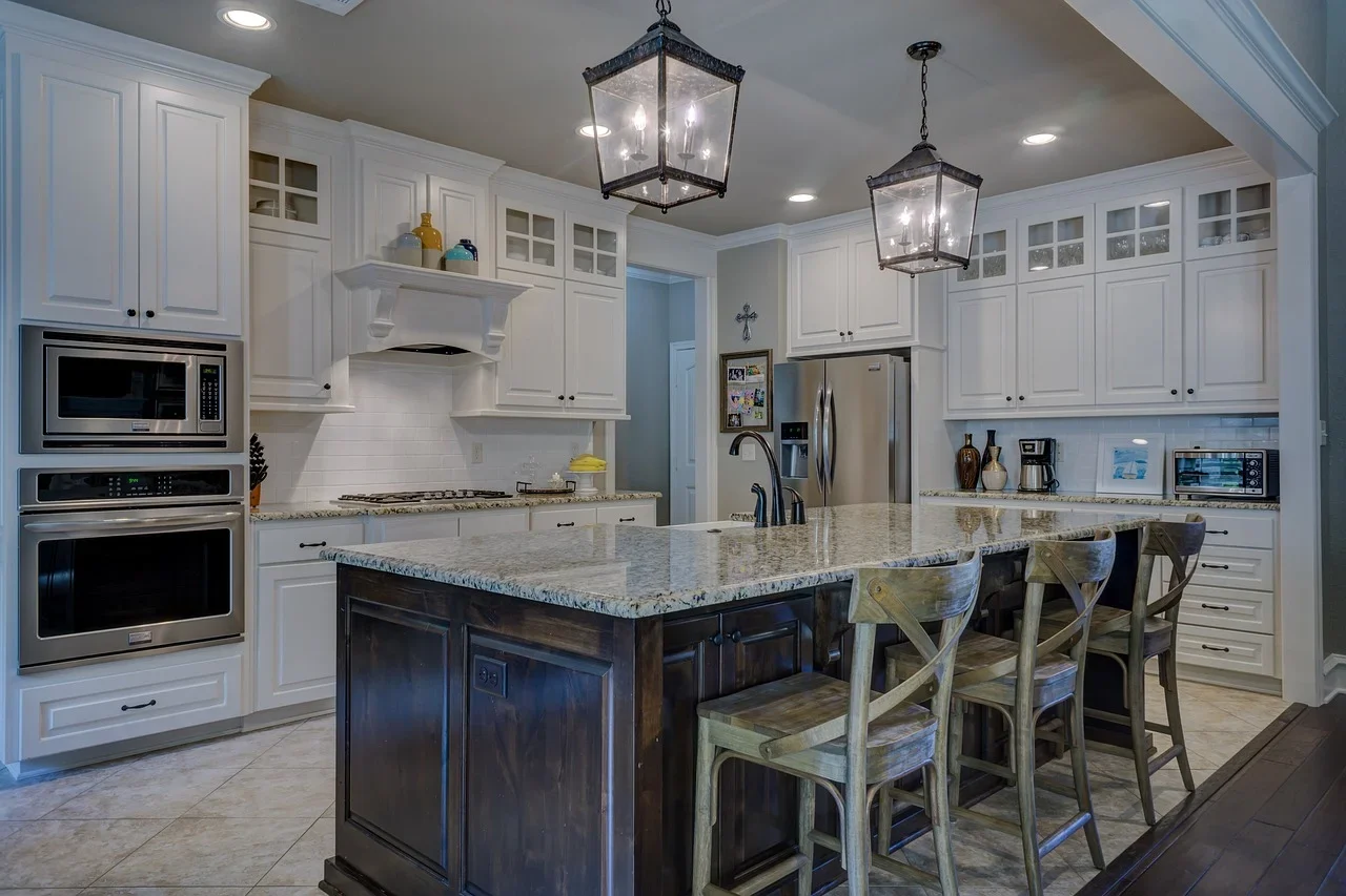 Modern kitchen with white cabinets, granite countertops, stainless steel appliances, and a central island with dark wood base and light granite top; four wooden chairs sit at the island. Two large pendant lights hang above the island, and recessed lighting is in the ceiling.