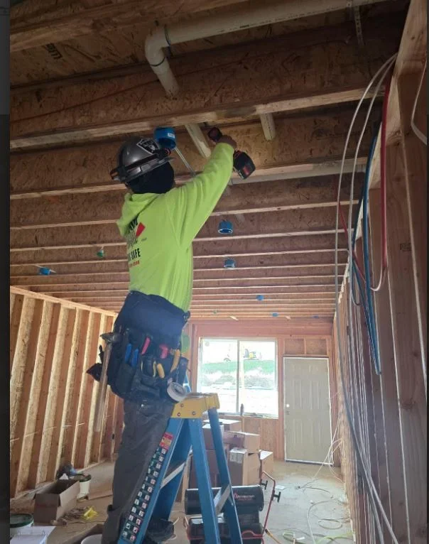 A construction worker installing electrical wiring on a ceiling inside a framed house. The worker is standing on a step ladder, wearing safety gear including a helmet and a tool belt.