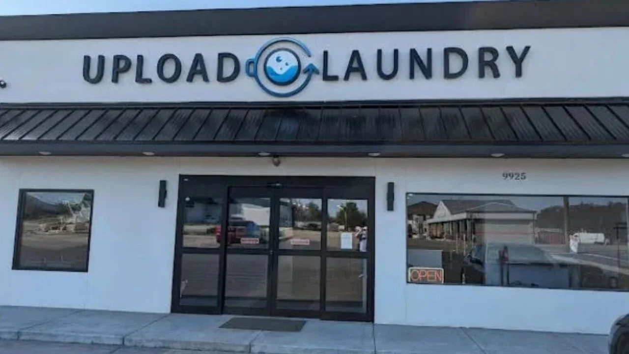 Front of a laundry business named Upload Laundry with a sign featuring a smiling washing machine logo, glass door entrance, and an illuminated 'Open' sign in the window.