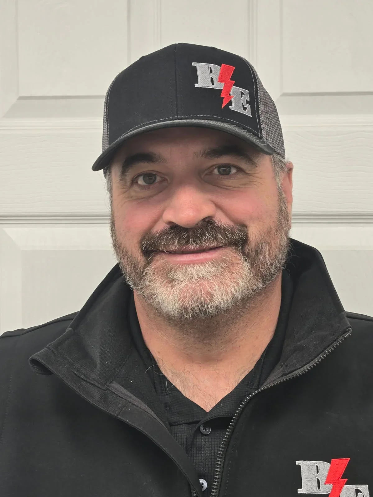 A smiling man with a beard wearing a black cap with a red lightning bolt and gray letters, black jacket, posing in front of a white garage door.