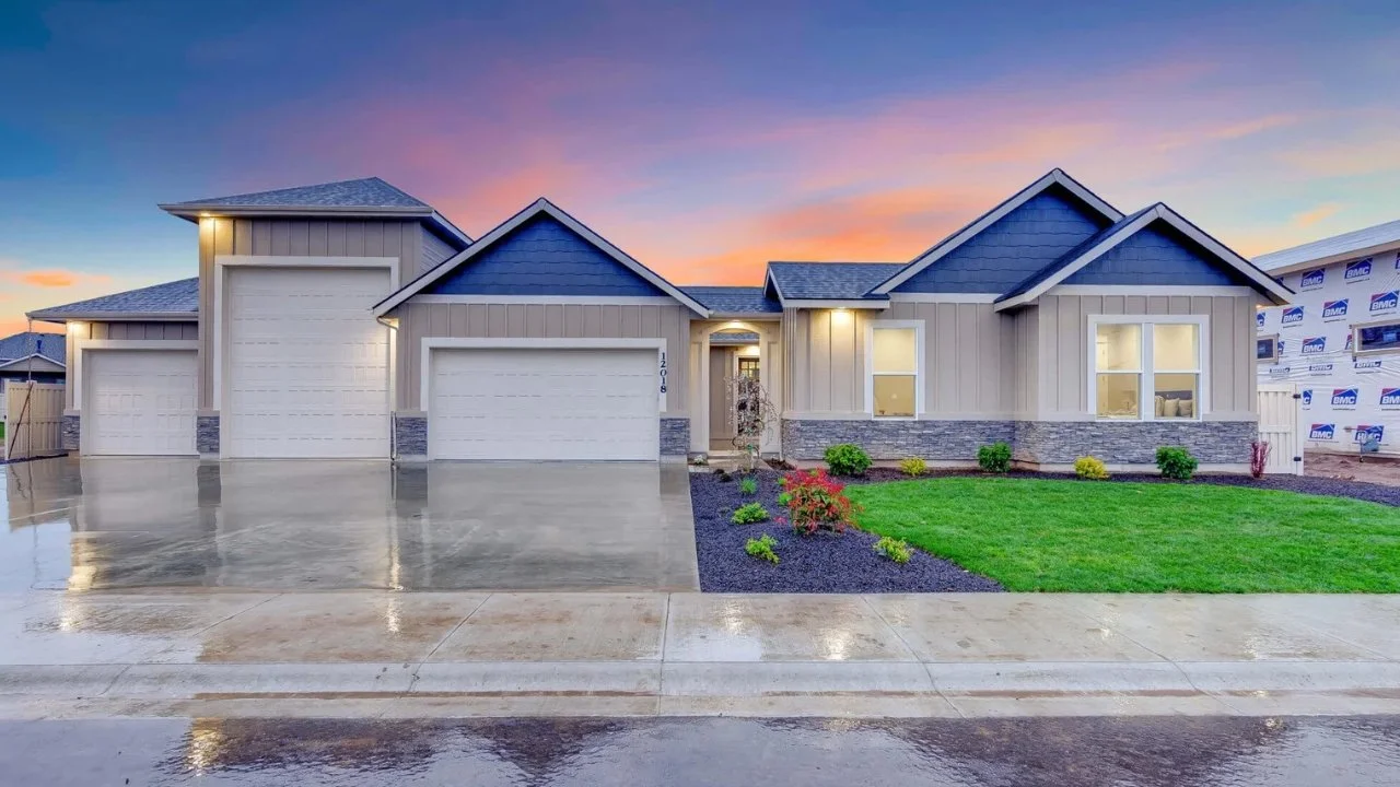 Newly built house with three garage doors, a small front yard with green grass and bushes, and a colorful sunset sky in the background.